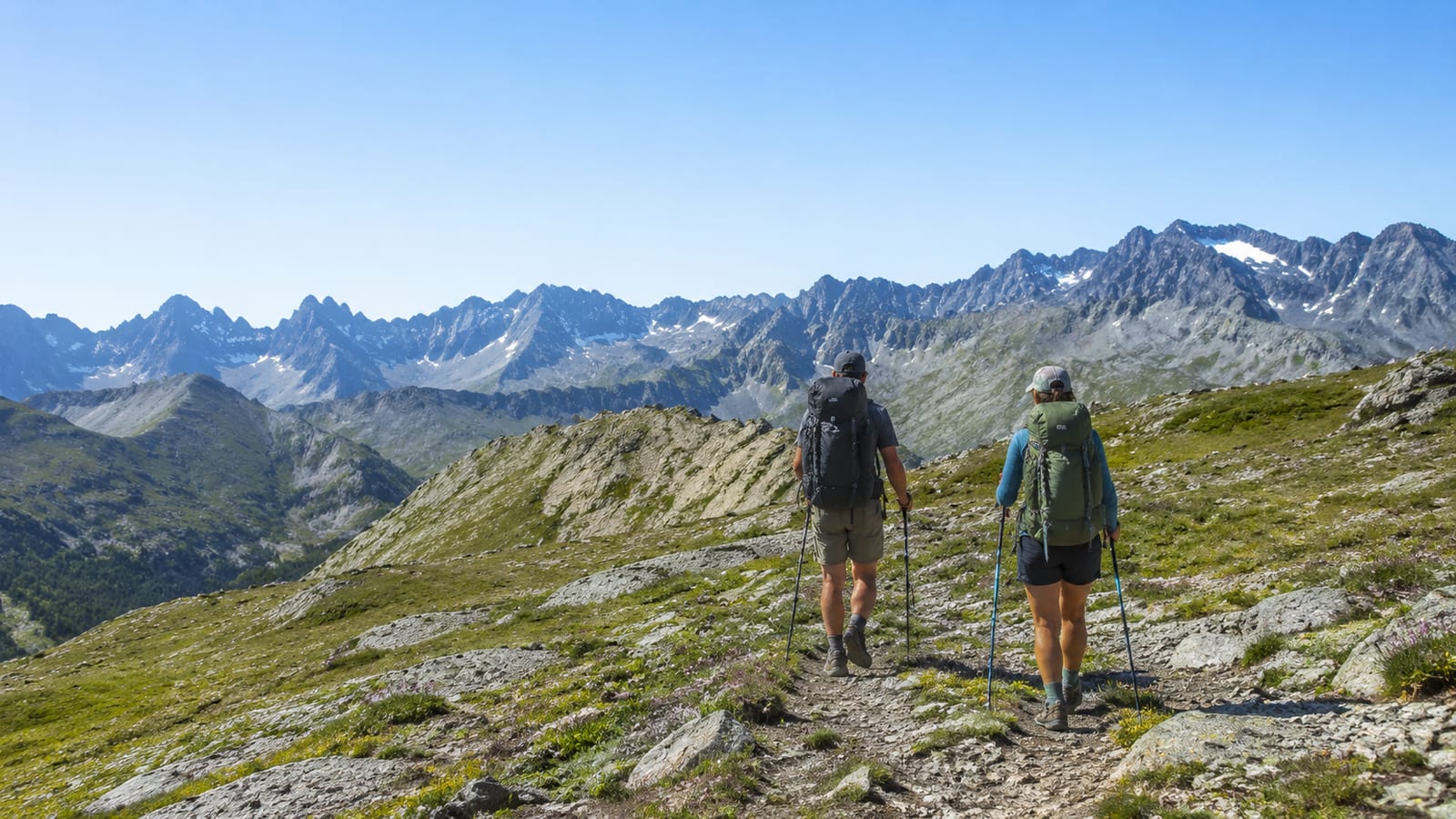 Two hikers cross a rocky alpine slope carrying daypacks and trekking poles
