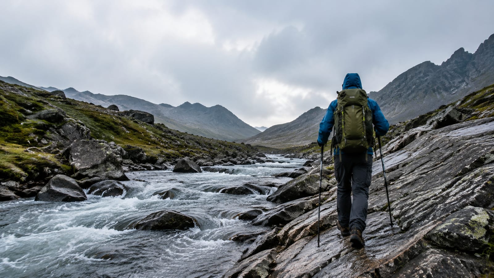 Hiker in rain shell navigates wet rocks beside a fast alpine stream
